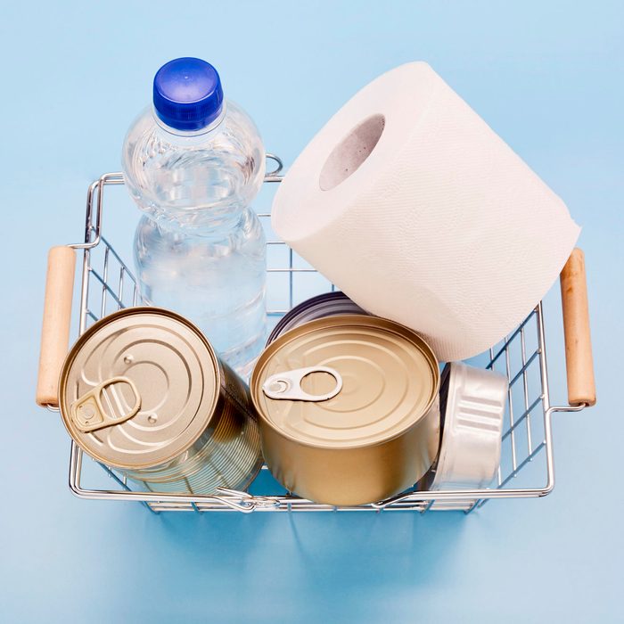 Still life of canned food, bottle of mineral water and toilet paper in shopping basket on blue background