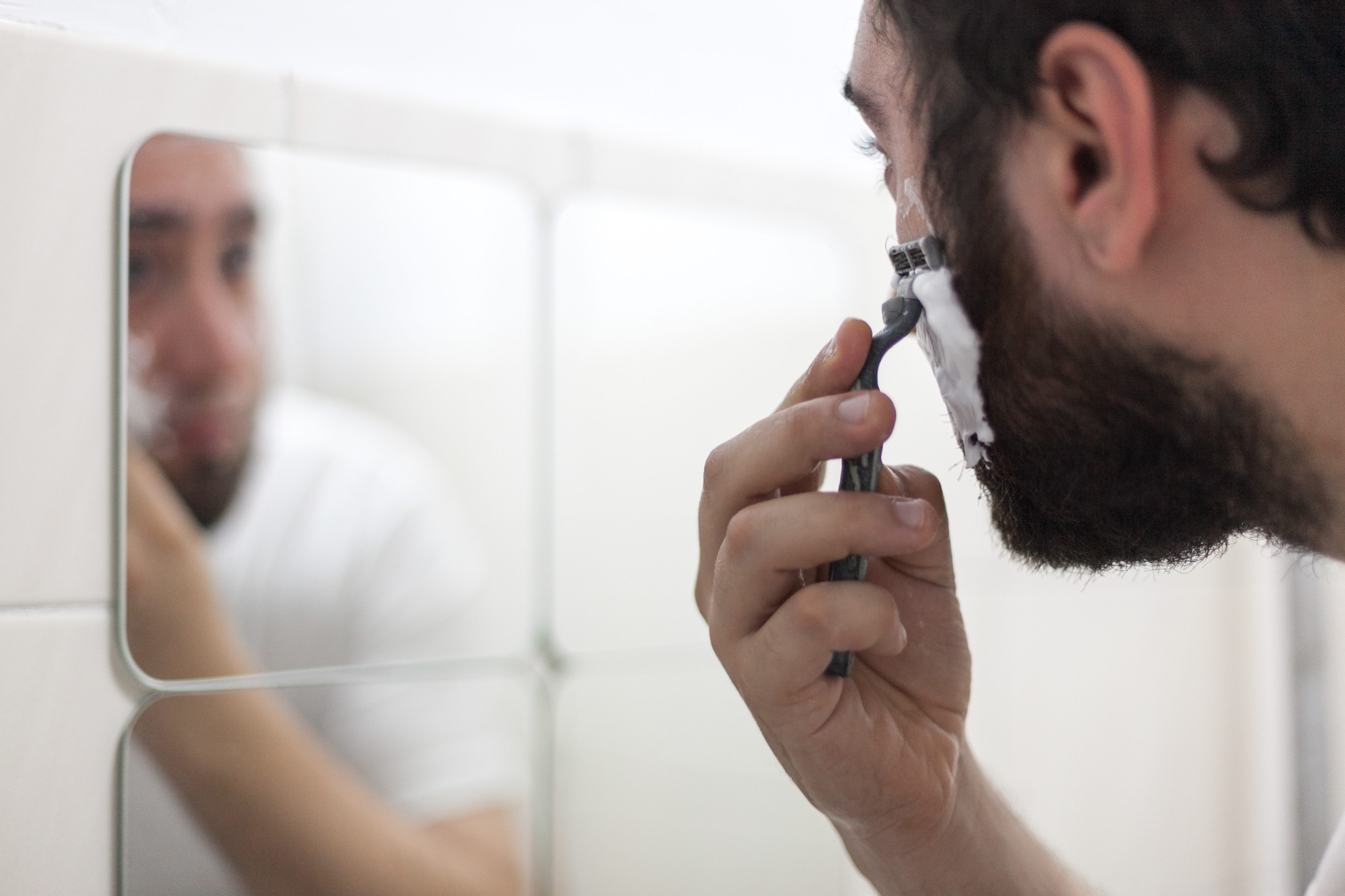 Man holding razor shaving face in front of mirror with foam on face