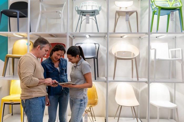 Friendly sales clerk helping adult couple look for furniture looking at a design on man's smartphone