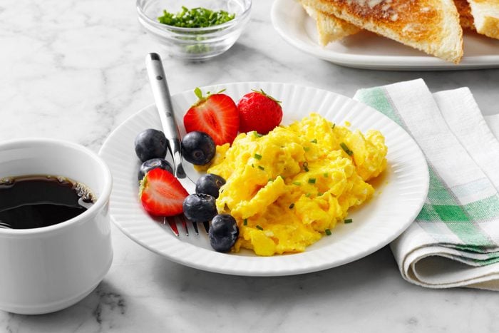 plate of scrambled eggs and berries on a white plate on marble kitchen counter with cup of coffee, toast, and fabric napkin