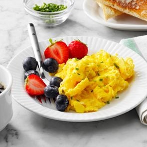 plate of scrambled eggs and berries on a white plate on marble kitchen counter with cup of coffee, toast, and fabric napkin