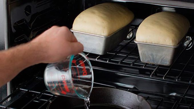 two loaves of bread sit in an open oven as a person pours water in a skillet below