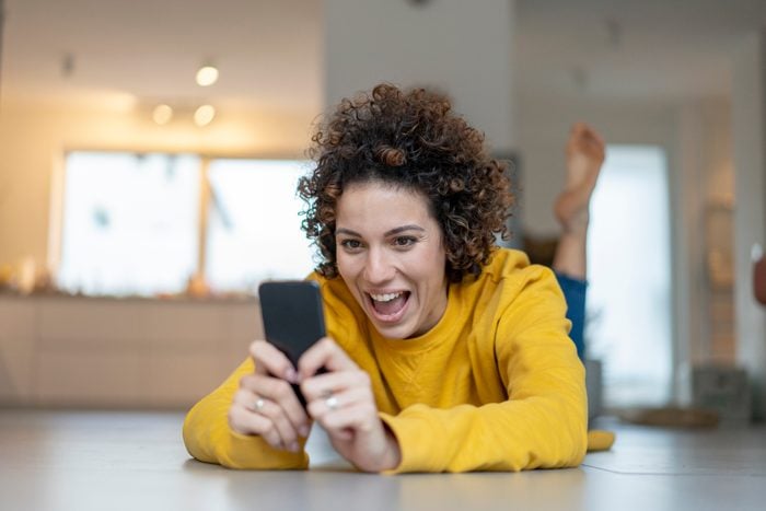 Excited woman lying on the floor at home using cell phone