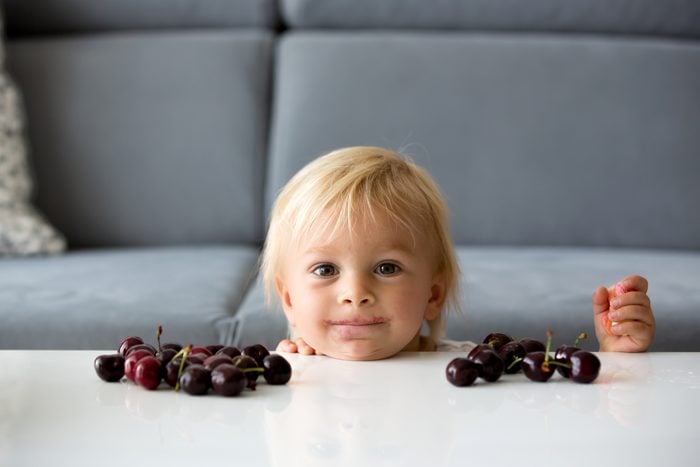 Sweet toddle boy, eating cherries, hiding behind a table, mischief look on his face