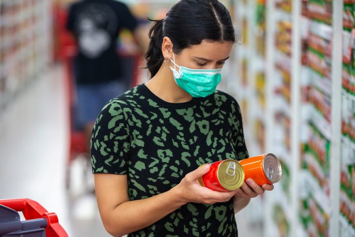 Alarmed female wears medical mask against coronavirus while grocery shopping in supermarket or store- health, safety and pandemic concept - young woman stockpiling food in fear of covid-19