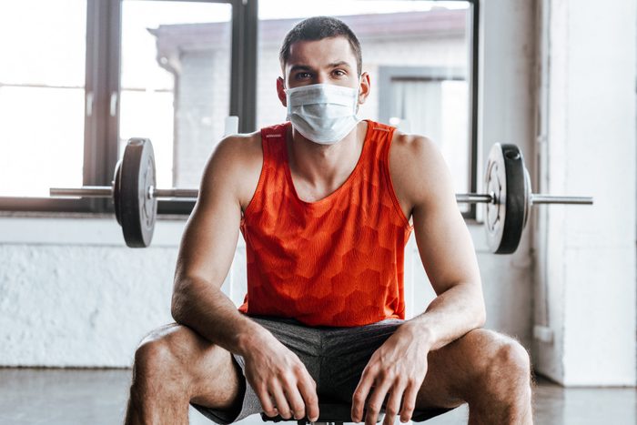 athletic sportsman in medical mask sitting near barbell in gym