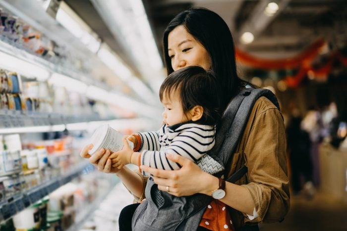 Mother carrying cute baby girl grocery shopping in supermarket and reading nutrition label on the package of fresh yoghurt
