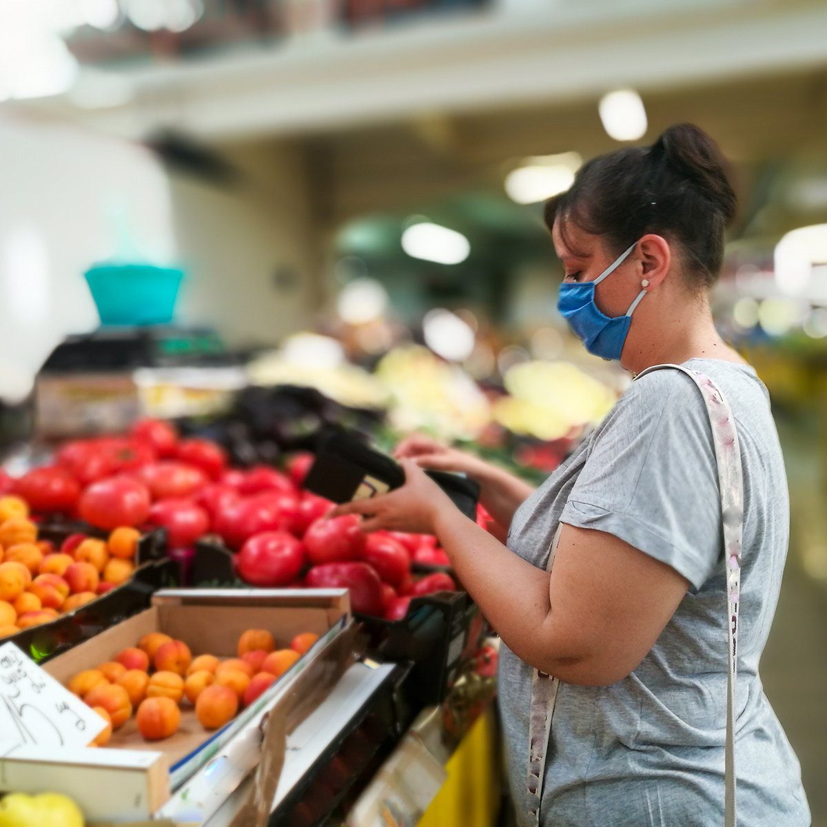 Selective focus color image depicting a caucasian woman in her 30s wearing a protective surgical face mask during the coronavirus (Covid-19) pandemic, in a bid to stop the spread of the virus. The woman is pushing shopping for fresh fruit and vegetables in her local market. Room for copy space.