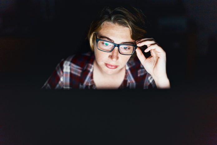 Serious young woman, lit by monitor against black, adjusts spectacles