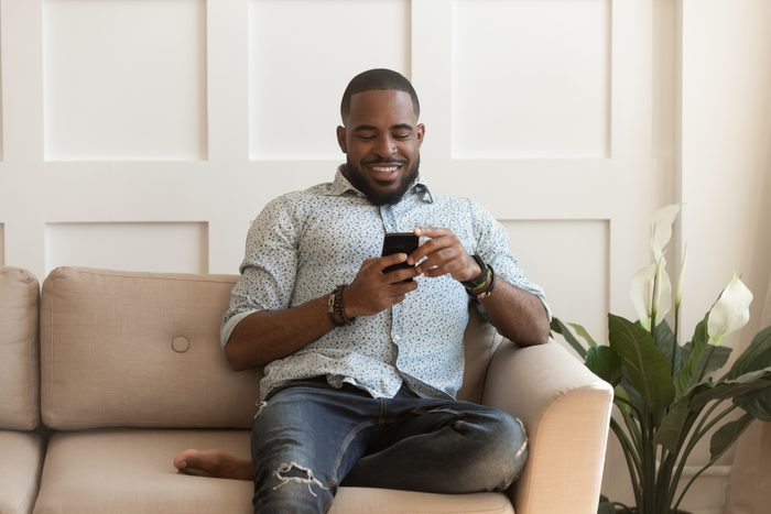 African guy sitting on couch chatting online using smartphone
