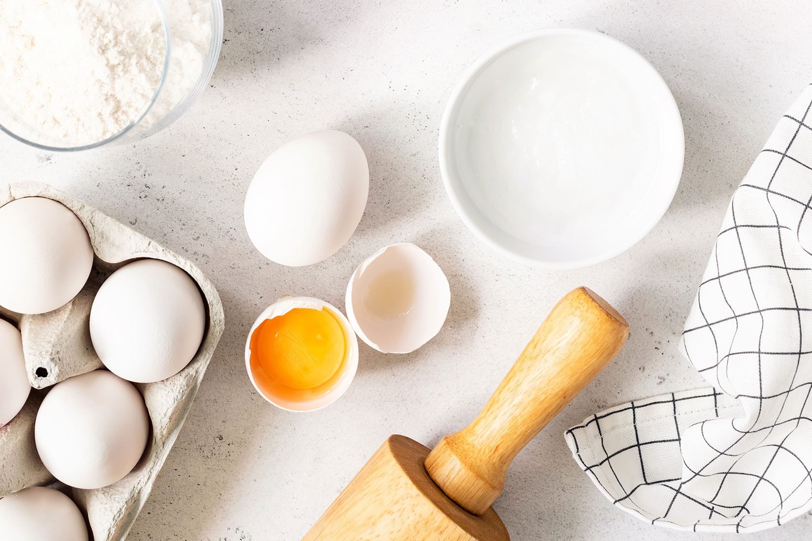 overhead view of eggs, rolling pin, baking soda, bowl on a white counter top