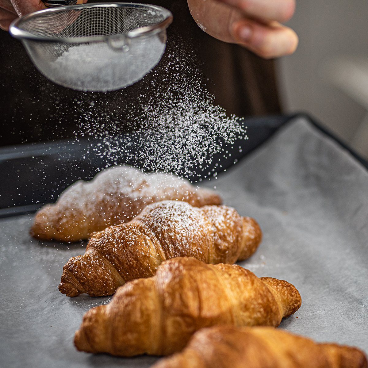 Close-up of chef's hand sprinkling powdered sugar on croissants with sieve in kitchen.