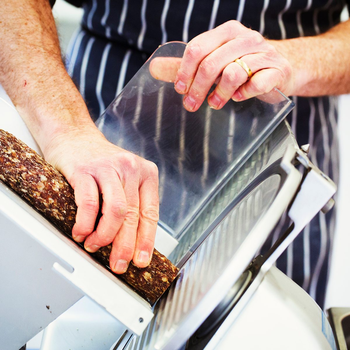 Close up of a butcher wearing a striped blue apron, slicing salami with a slicer.