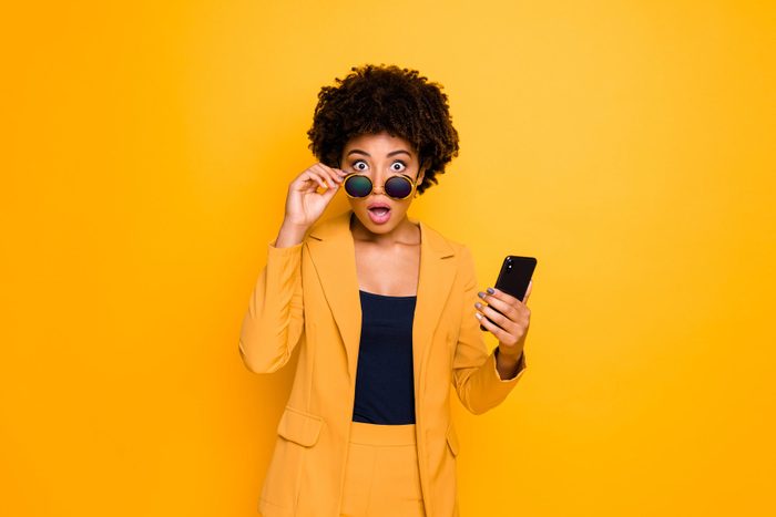 Portrait of shocked afro american girl use smartphone read news on social network account get notification about dislike scream omg wear stylish fashionable clothes isolated yellow color background