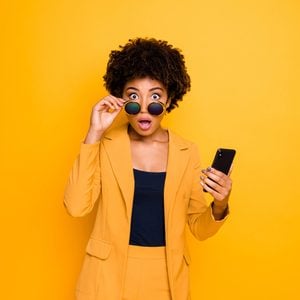 Portrait of shocked afro american girl use smartphone read news on social network account get notification about dislike scream omg wear stylish fashionable clothes isolated yellow color background