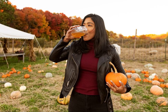 young woman drinking apple cider and holding a pumpkin at the pumpkin patch during autumn