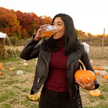 young woman drinking apple cider and holding a pumpkin at the pumpkin patch during autumn