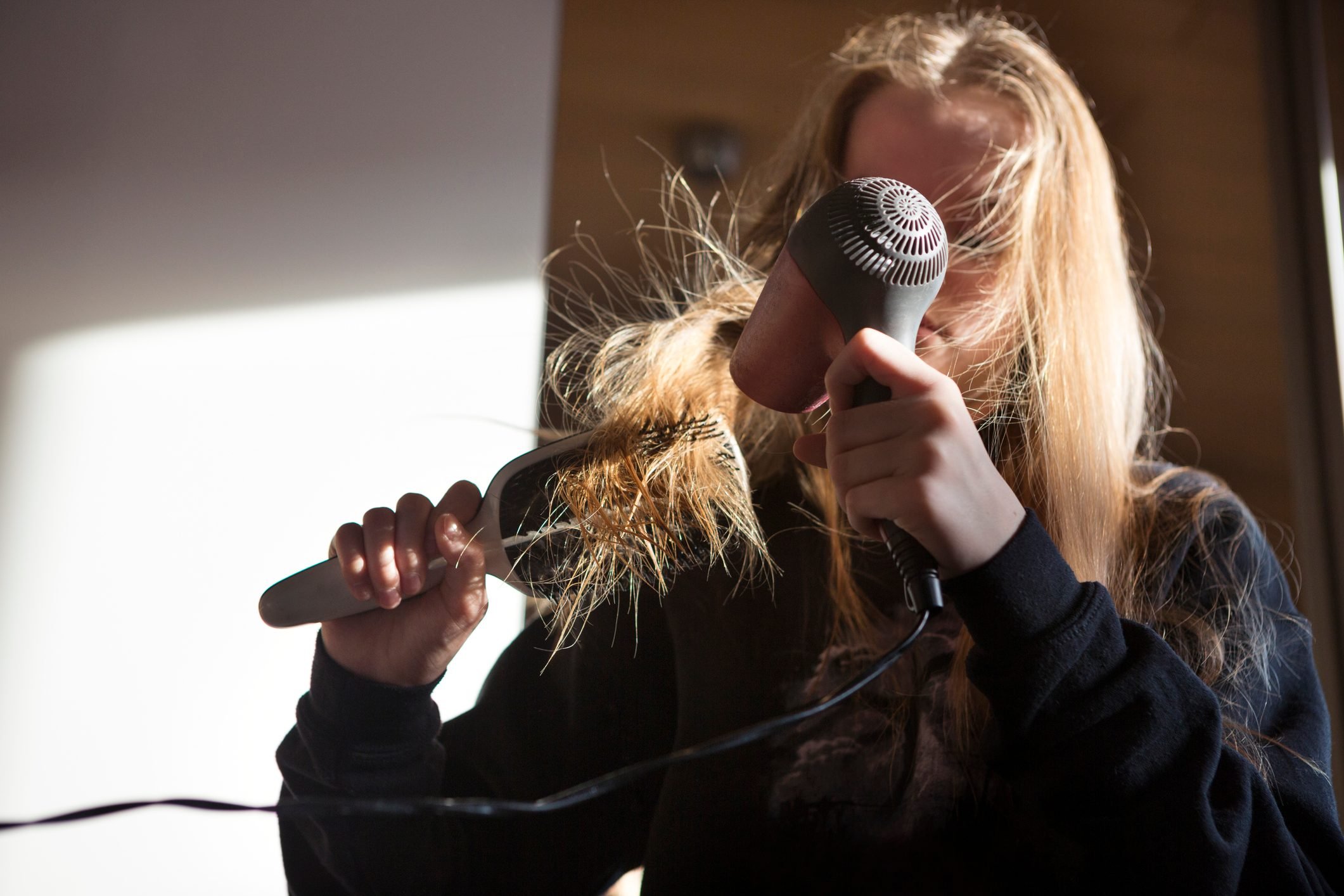 Girl (12-13) blow drying her long hair with an electric hairdryer in a bedroom