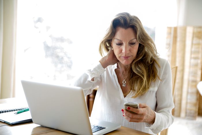 Mature woman using laptop whilst reading smartphone text at home