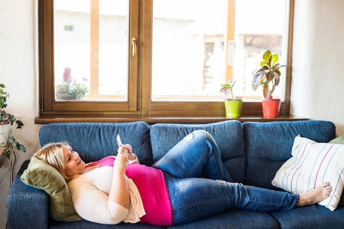 Attractive overweight woman at home lying on couch, holding smart phone, texting.