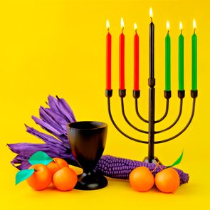 Contemporary Kwanzaa display with corn, apples, candles, and chalice on yellow background.
