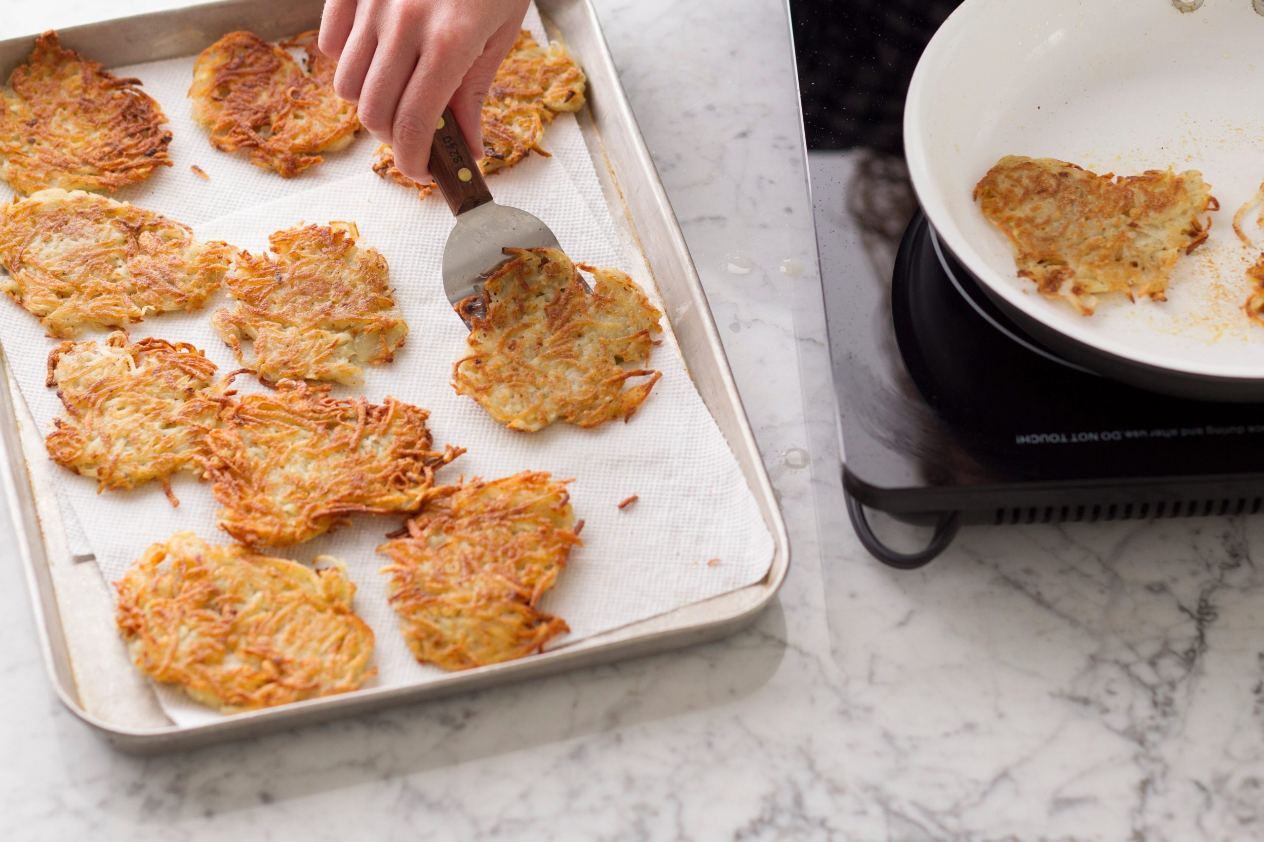 Metal spatula depositing potato pancakes from the skillet to a lined baking sheet