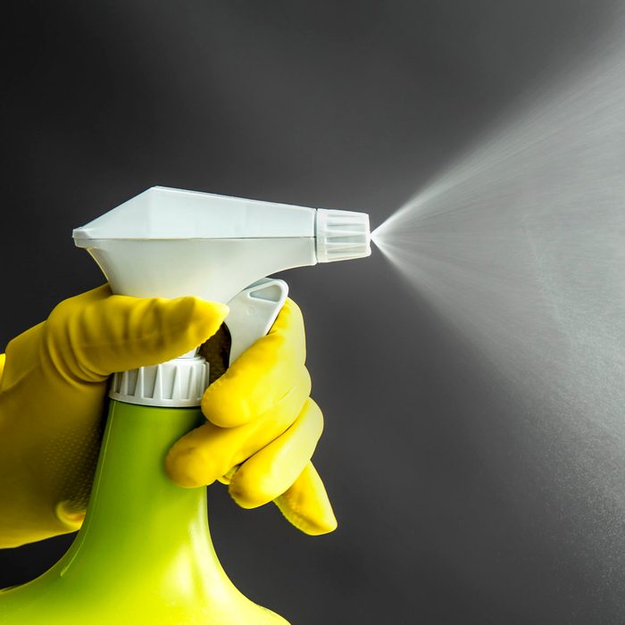 Woman wearing yellow rubber gloves using green spray bottle and spraying liquid mist in air, cool lighting effect. Lot of copy space.