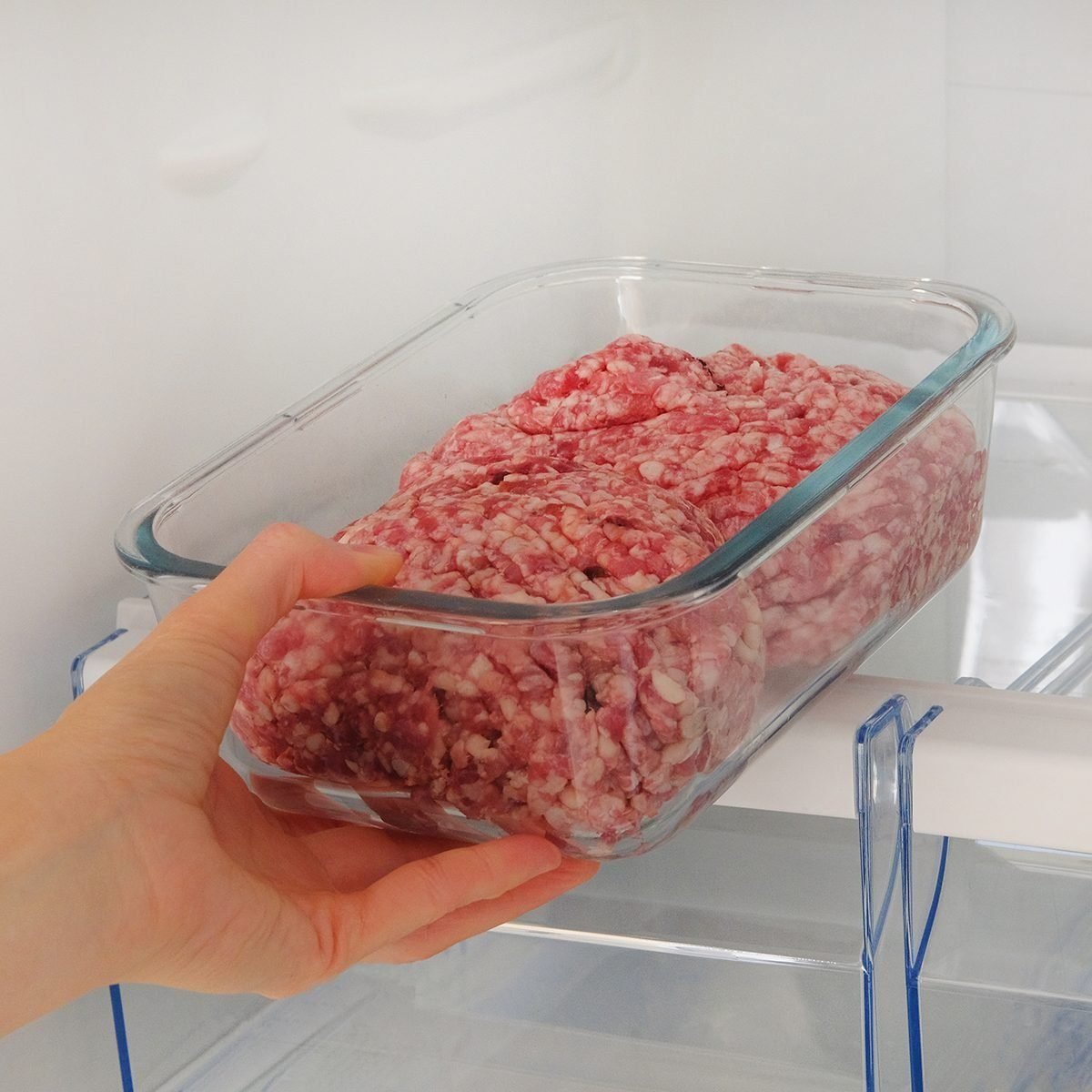 Minced raw meat in glass container is on shelf in open fridge.