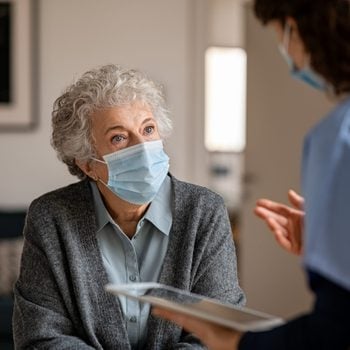 Senior woman wearing safety protective mask at home and talking to nurse. Back view of young doctor visiting old woman for routine health checkup during covid-19, coronavirus and flu outbreak.
