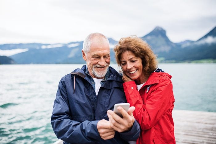 Cheerful senior couple tourist standing by lake in nature on holiday, using smartphone.