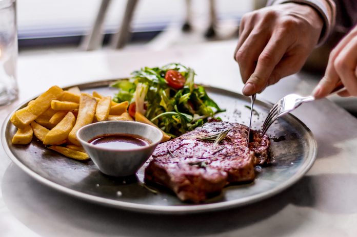 Close-Up Of Hands Cutting Into Steak Served On Plate With Fries