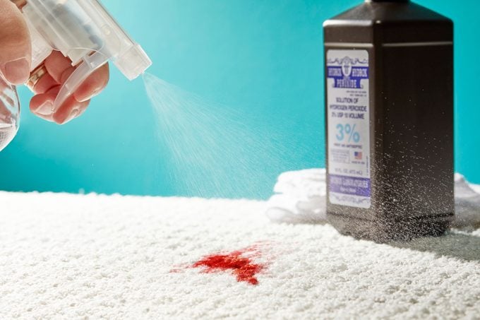 Blood stain on a white carpet with hand spraying a spray bottle and bottle of 3% hydrogen peroxide and white cleaning cloths nearby
