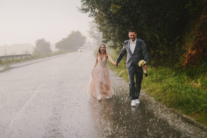 Just married couple holding hands and walking on rain. Walking in wet ceremonial clothes on driveway. Smiling and looking at camera.