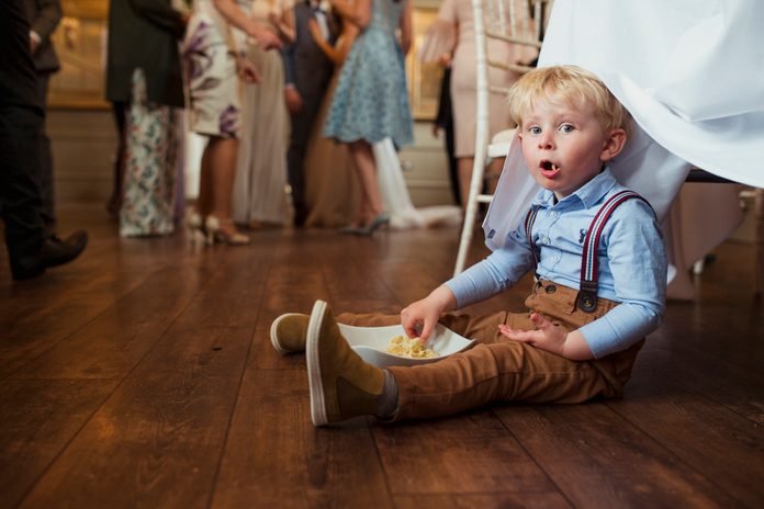 Young boy sitting under a table at a wedding reception with a bowl of snacks.