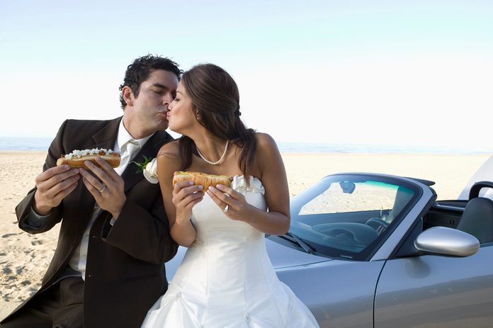 newlyweds kissing while holding hotdogs and posing by thier convertible on the beach