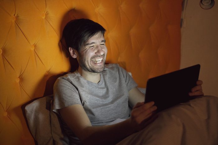 Young laughing man using tablet computer for surfing social media lying in bed at home before sleeping