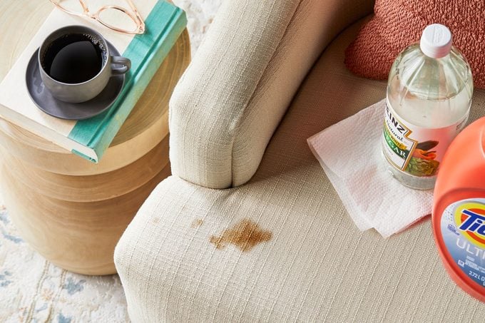 coffee stain on a couch. cup of coffee and book on side table nearby. cleaning supplies also nearby