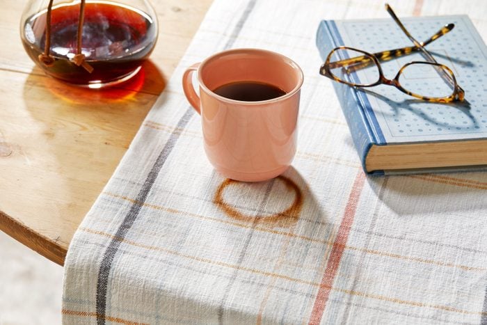 cup of coffee and a ring stain on fabric runner on a kitchen table. carafe and book nearby on the table