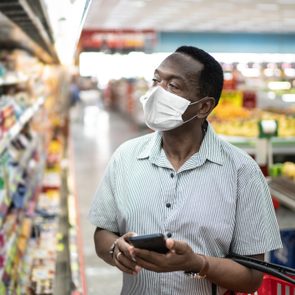 Mature man using mobile and choosing products in supermarket - using face mask