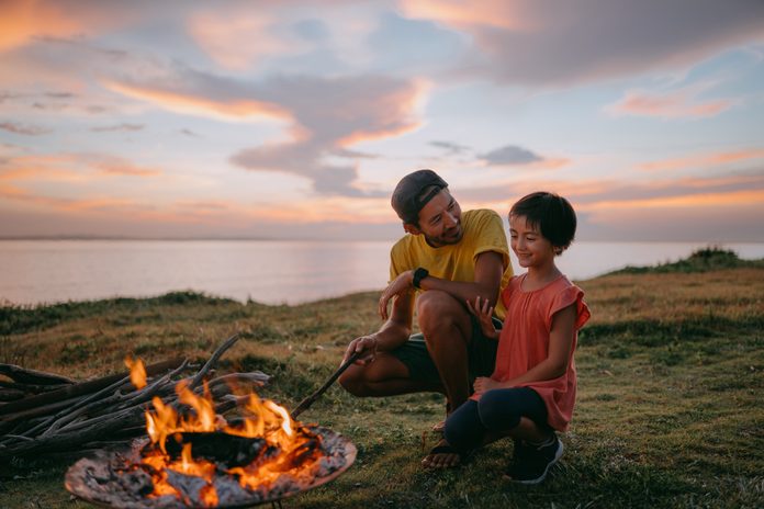 Father and daughter enjoying campfire at seaside campsite at sunset