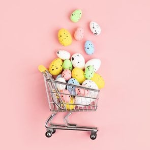 toy grocery cart on it's side on pink background with candy easter eggs pouring out of the basket