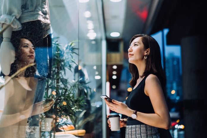 Cheerful young Asian woman holding a cup of coffee, checking her smartphone while standing outside a boutique looking at shop window in the evening in the city