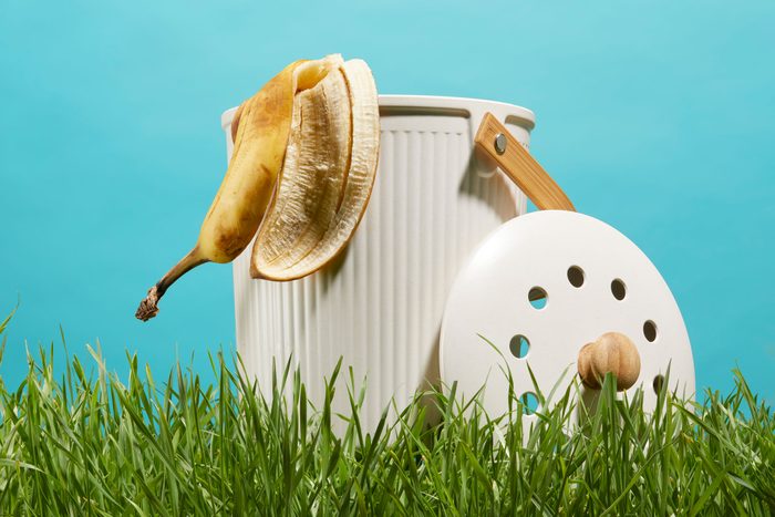 small white compost bin in the grass with turquoise background with a banana peel hanging out over the top edge