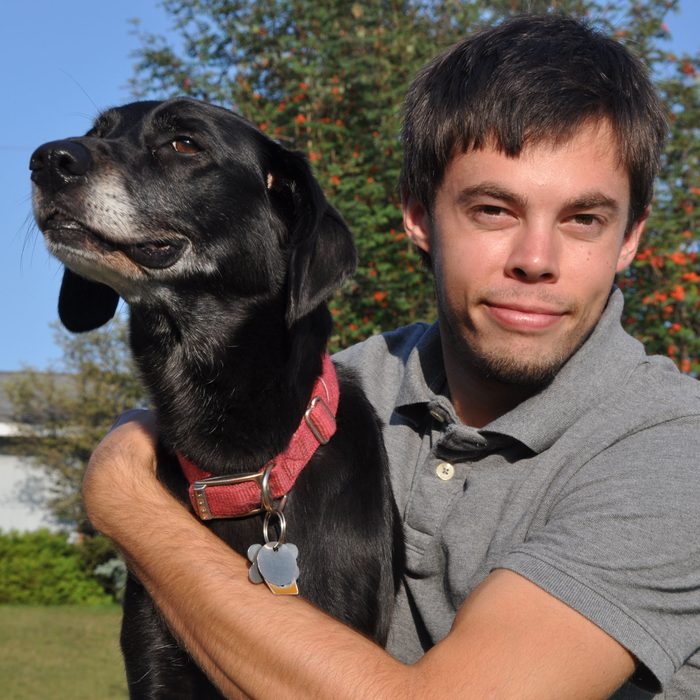a young man sits with his arm around a black dog