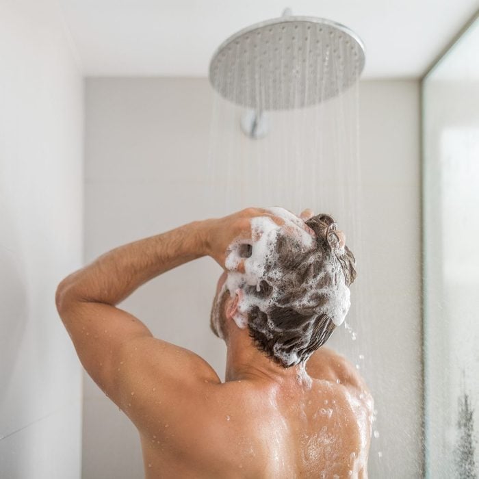 Man taking a shower washing hair under water falling from rain showerhead in luxury walk-in bath. Showering young person at home lifestyle. Body care morning routine in sunlight