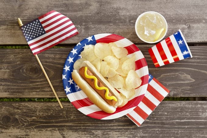 fourth of july still life: overhead view of small american flag, flag plate with hot dog and chips, and flag cups and napkin on a wood table background