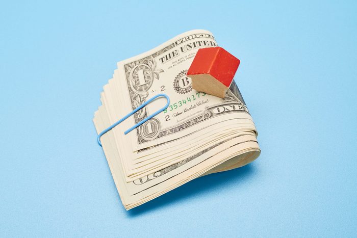 bundle of folded money held with a paper clip and small, wooden house on blue background