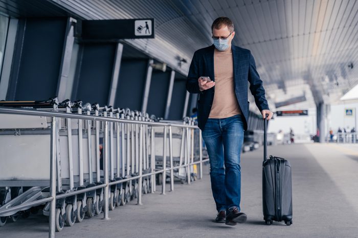 Male passenger walks in airport, holds suitcase, checks newsfeed on smartphone, wears disposable medical mask during pandemic coronavirus, tries to be safe during disease virus. Respiratory symptoms