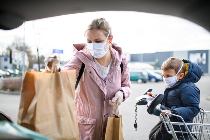 Mother and small son with face mask outdoors putting grocery bags in car.