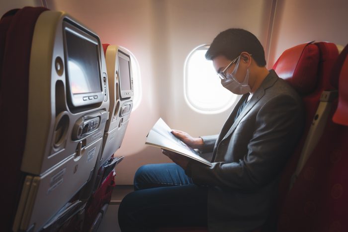 Young asian businessman wearing protective face mask with suit sitting on airplane seat while reading magazine due to Coronavirus or COVID-19 outbreak situation in all of landmass in the world